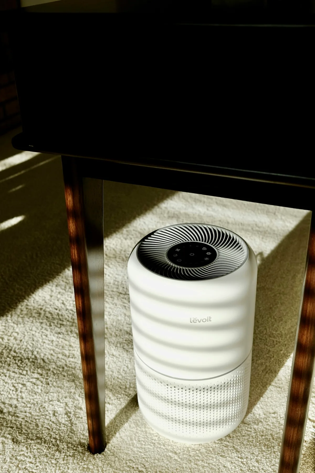 A white air purifier beneath a desk beside dark furniture